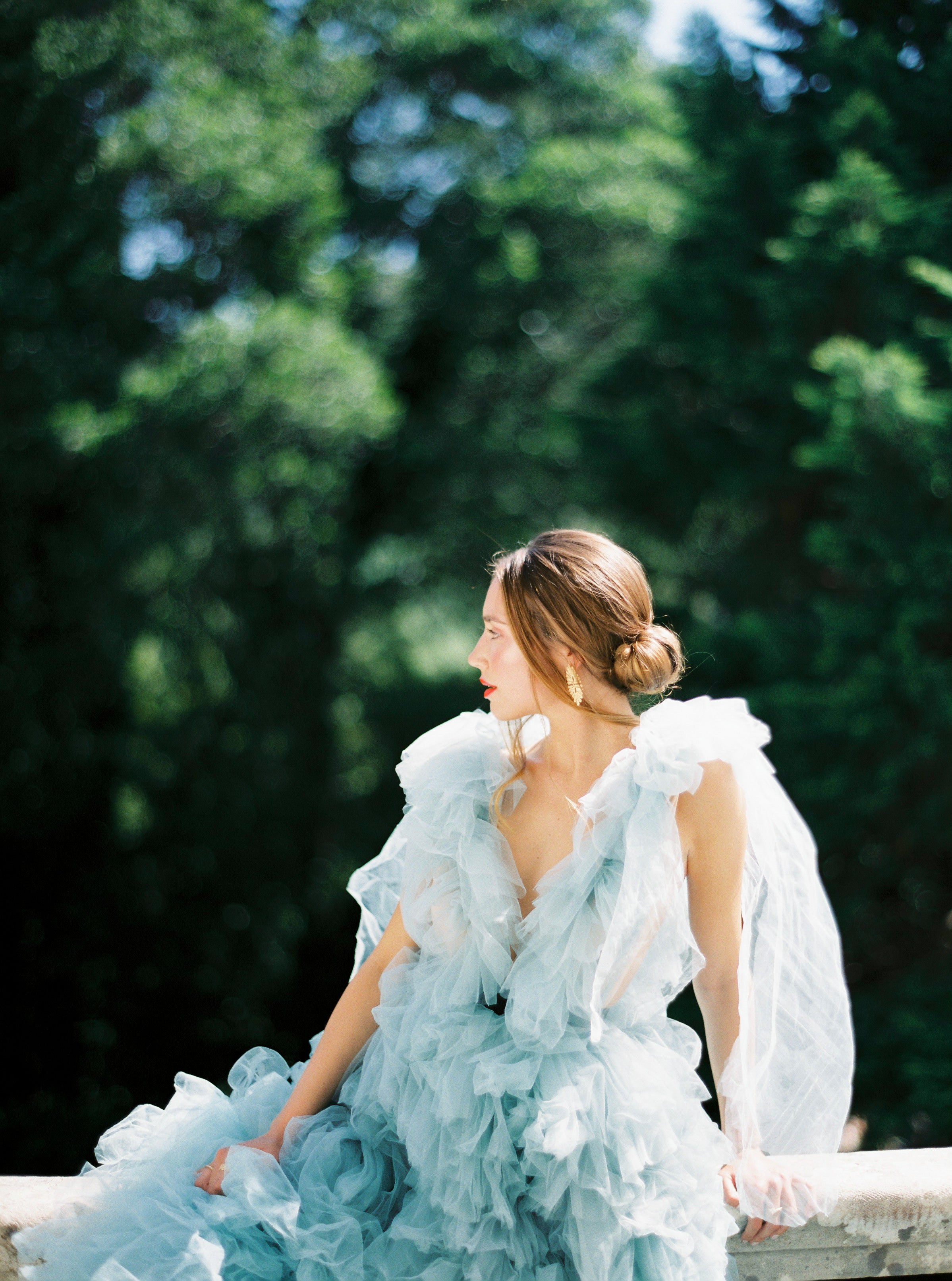 A vintage-inspired bride in a blue tulle dress sitting on a railing, adorned with elegant Cremilde Bispo Jewellery's Guipure GP lace artistry.