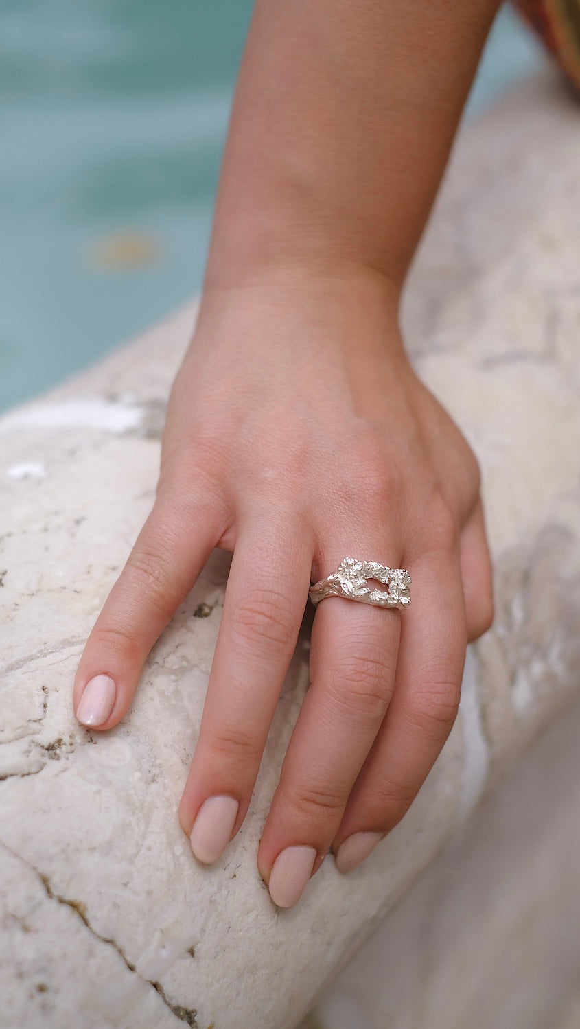 A woman's hand holding the Cremilde Bispo Jewellery Glade ring next to a pool surrounded by floral foliage.