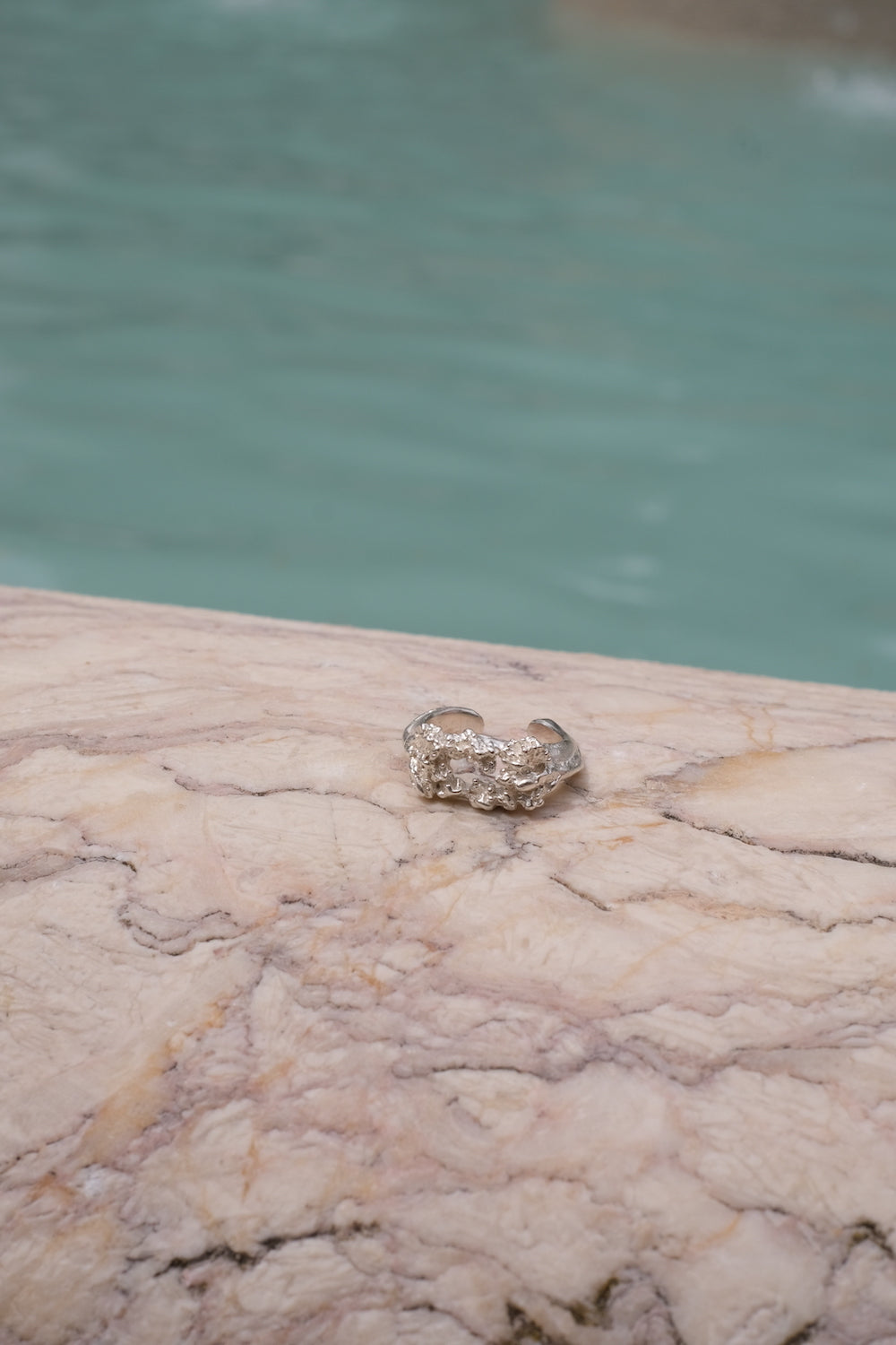 A handcrafted Glade ring by Cremilde Bispo Jewellery adorns a marble slab nestled in a glade beside a shimmering pool, surrounded by delicate floral foliage.