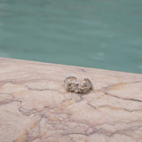 A handcrafted Glade ring by Cremilde Bispo Jewellery adorns a marble slab nestled in a glade beside a shimmering pool, surrounded by delicate floral foliage.