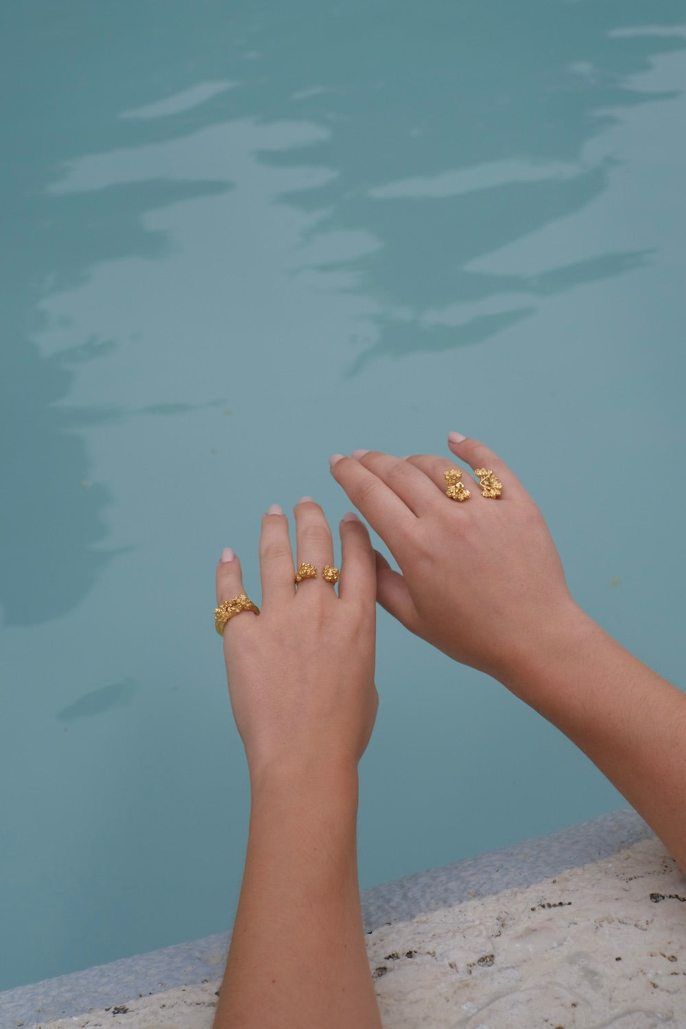 A woman's hands adorned with a Cremilde Bispo Jewellery's handcrafted jewelry collection, featuring The Glade ring GP, are elegantly displayed next to a pool.