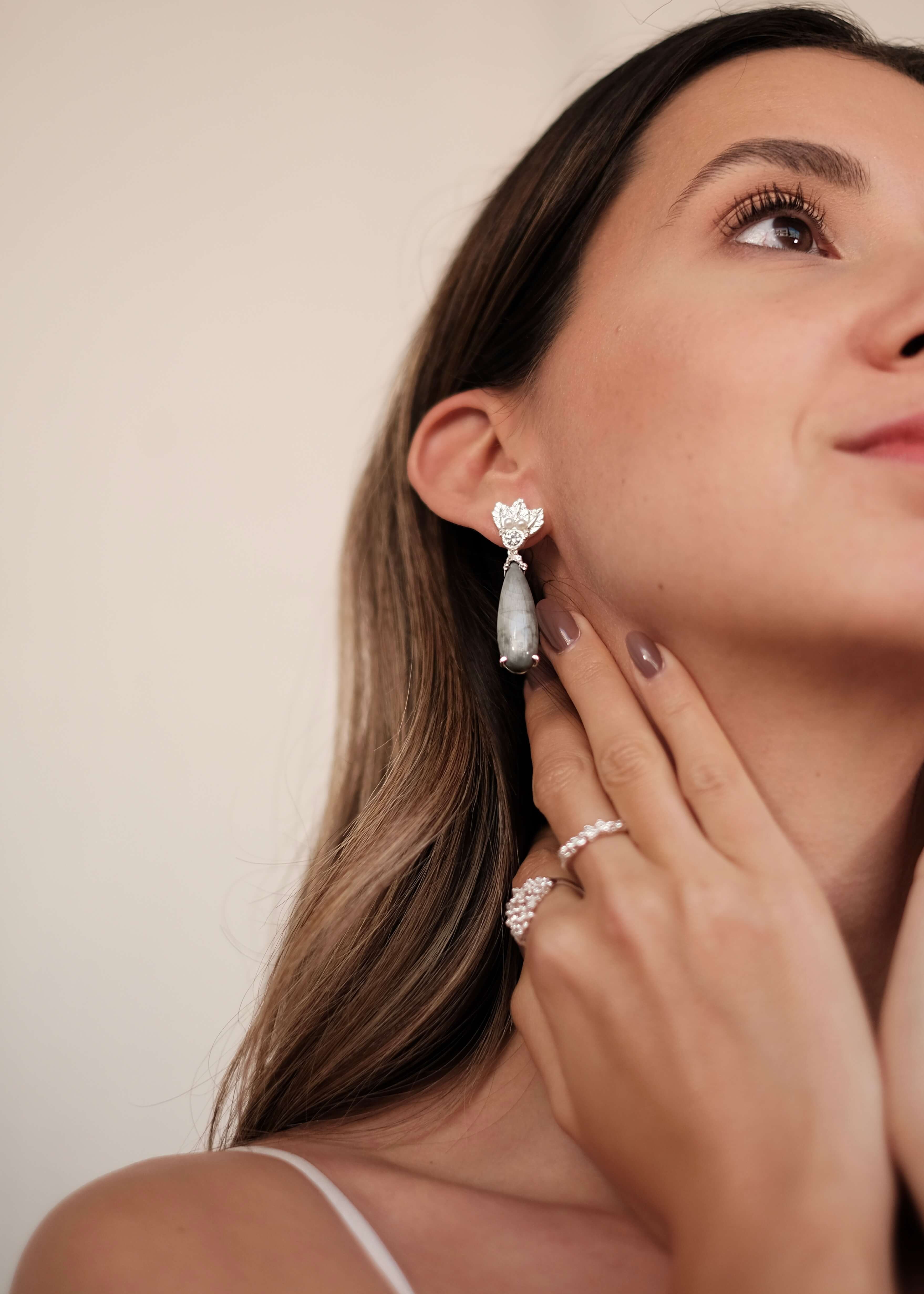 A woman wearing a pair of The Muse III Toed eye earrings with natural drop-shaped gemstones by Cremilde Bispo Jewellery.