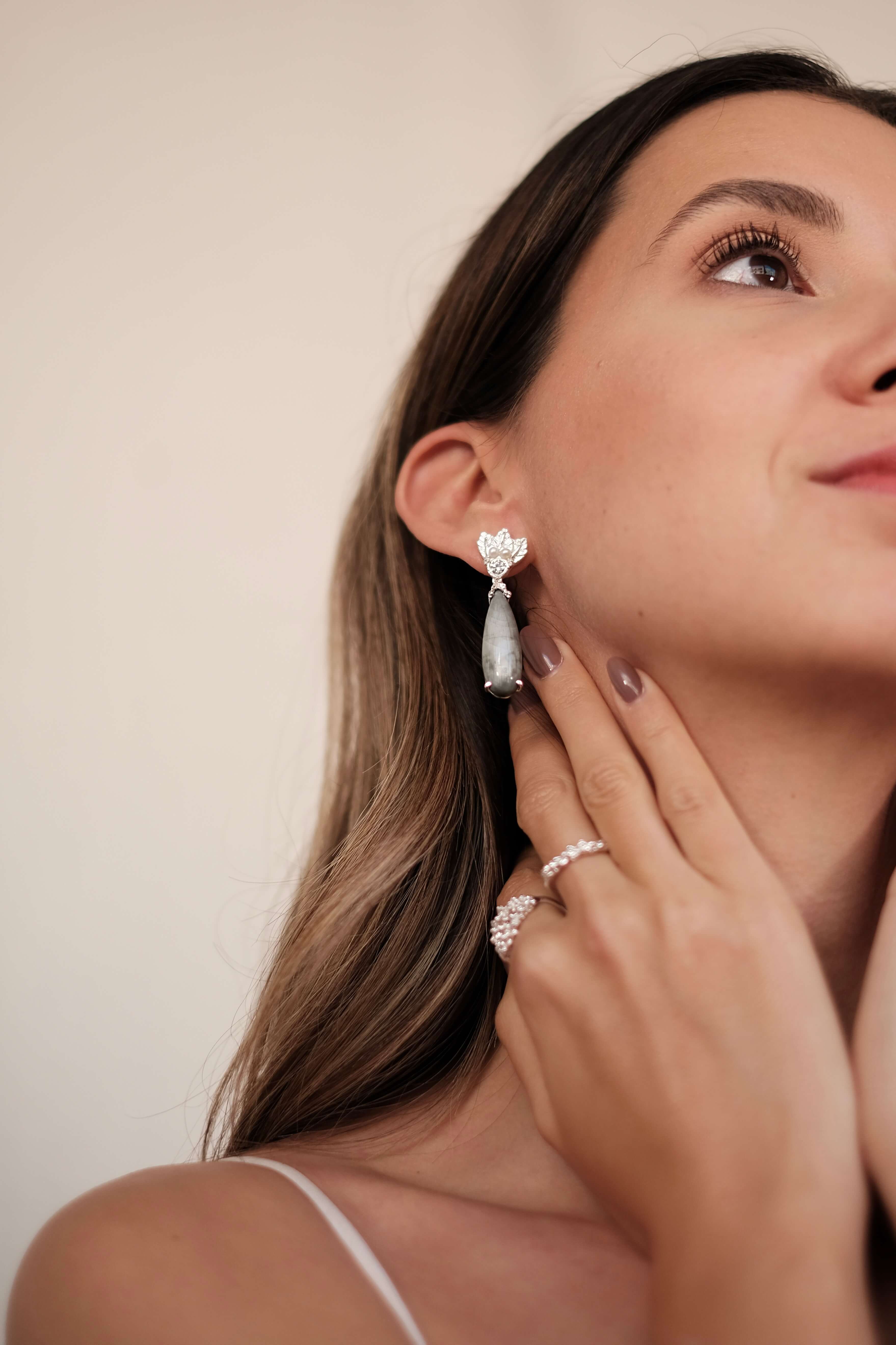 A woman wearing a pair of The Muse III Toed eye earrings with natural drop-shaped gemstones by Cremilde Bispo Jewellery.