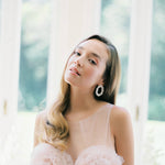 A bride in a pink dress gracefully posing in front of a window, showcasing the refined elegance of Nature's beauty, perfectly complemented by her Cremilde Bispo Jewellery Garden's Delight Earrings, in Sterling silver.