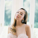 A bride in a pink dress gracefully posing in front of a window, showcasing the refined elegance of Nature's beauty, perfectly complemented by her Cremilde Bispo Jewellery Garden's Delight Earrings, in Sterling silver.