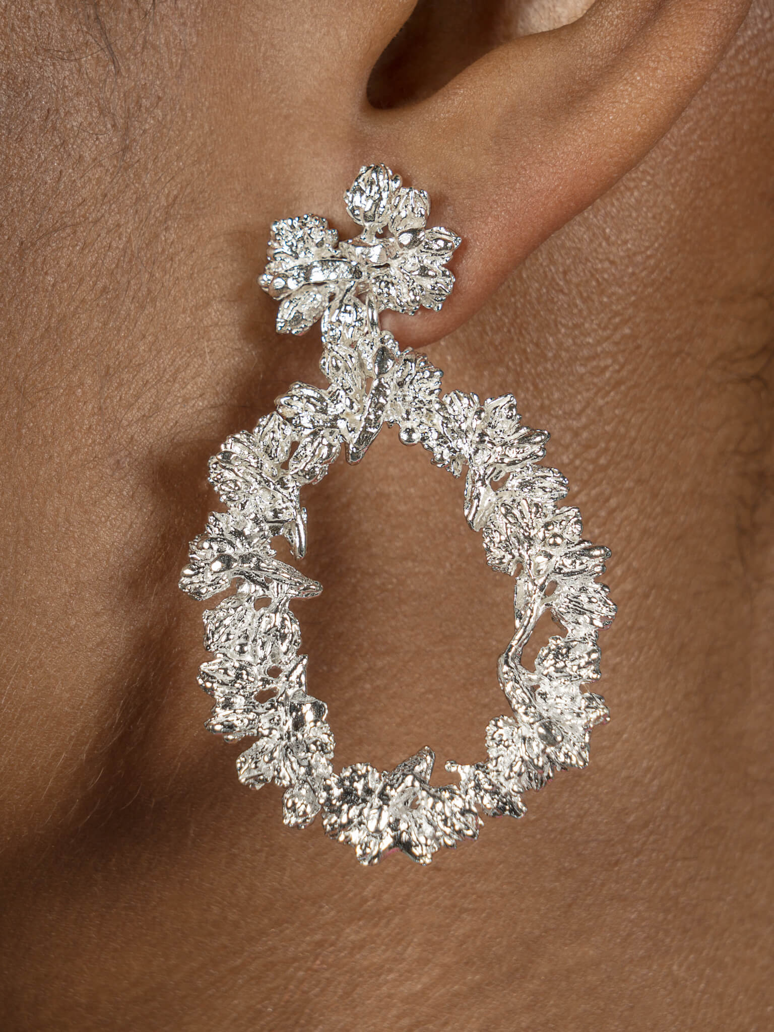 A woman's ear adorned with the Cremilde Bispo Jewellery Garden's Delight statement earrings. Handcrafted from sterling silver, these earrings feature meticulously hand-sculpted floral accents and an elegant drop-shaped design that gently sways with grace.