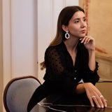 A sophisticated woman wearing the Cremilde Bispo Jewellery Garden's Delight statement earrings in sterling silver, seated at an opulently decorated table in a luxurious room.