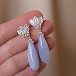A hand holding a pair of The Muse III Chalcedony earrings, adorned with .925 silver accents by Cremilde Bispo Jewellery.