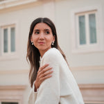 A woman wearing a white sweater and Cremilde Bispo Jewellery's Pearly Forest Earrings, standing on a balcony.