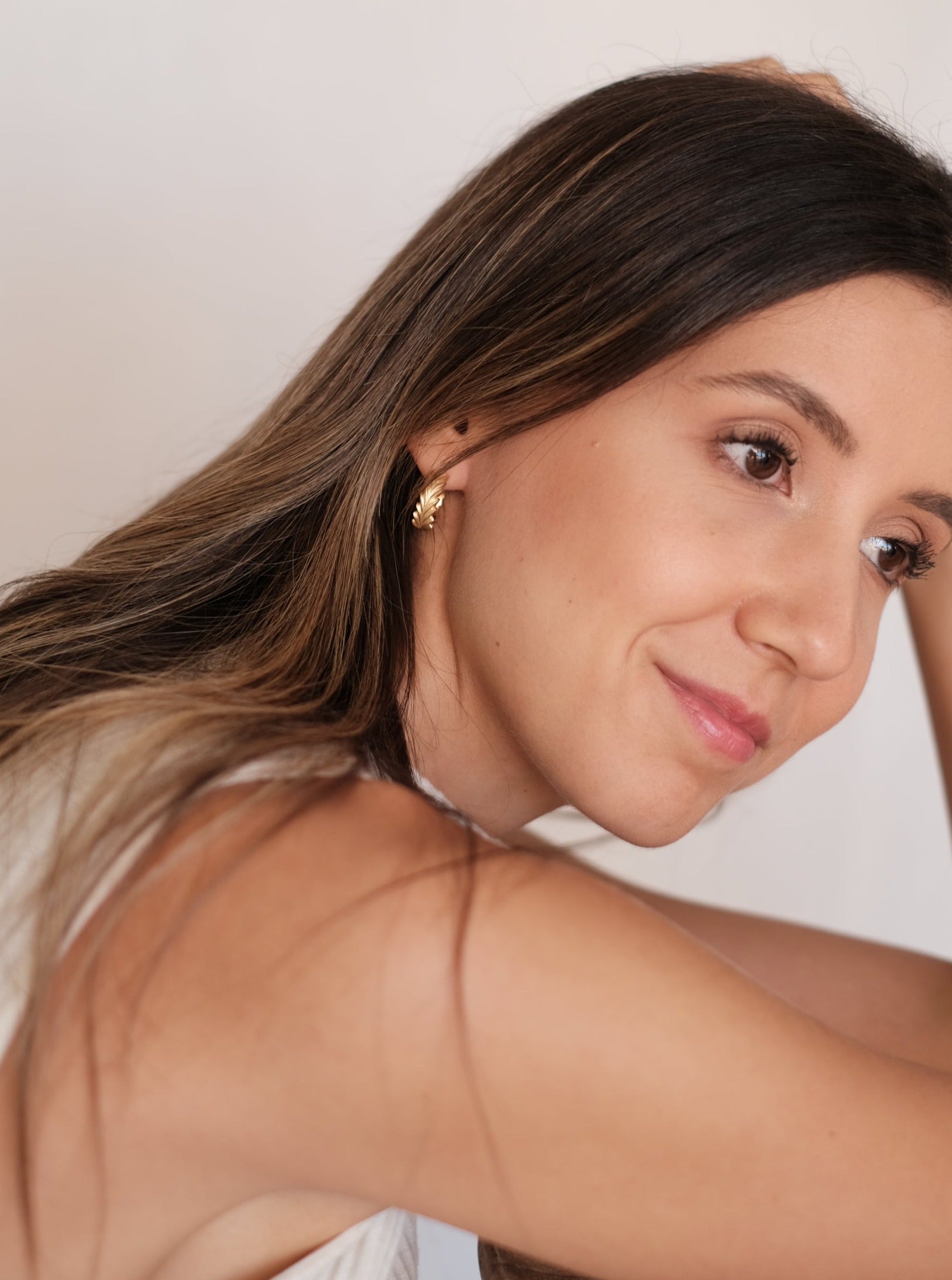 A woman showcasing timeless elegance leans against a wall, her hand gracefully placed on her head. The image exudes exclusive touch and is complemented by the Cremilde Bispo Jewellery's Essential Leaf Hoops GP.