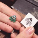 A person is holding The Muse I, a handcrafted jewelry piece by Cremilde Bispo Jewellery, with a leaf on it.