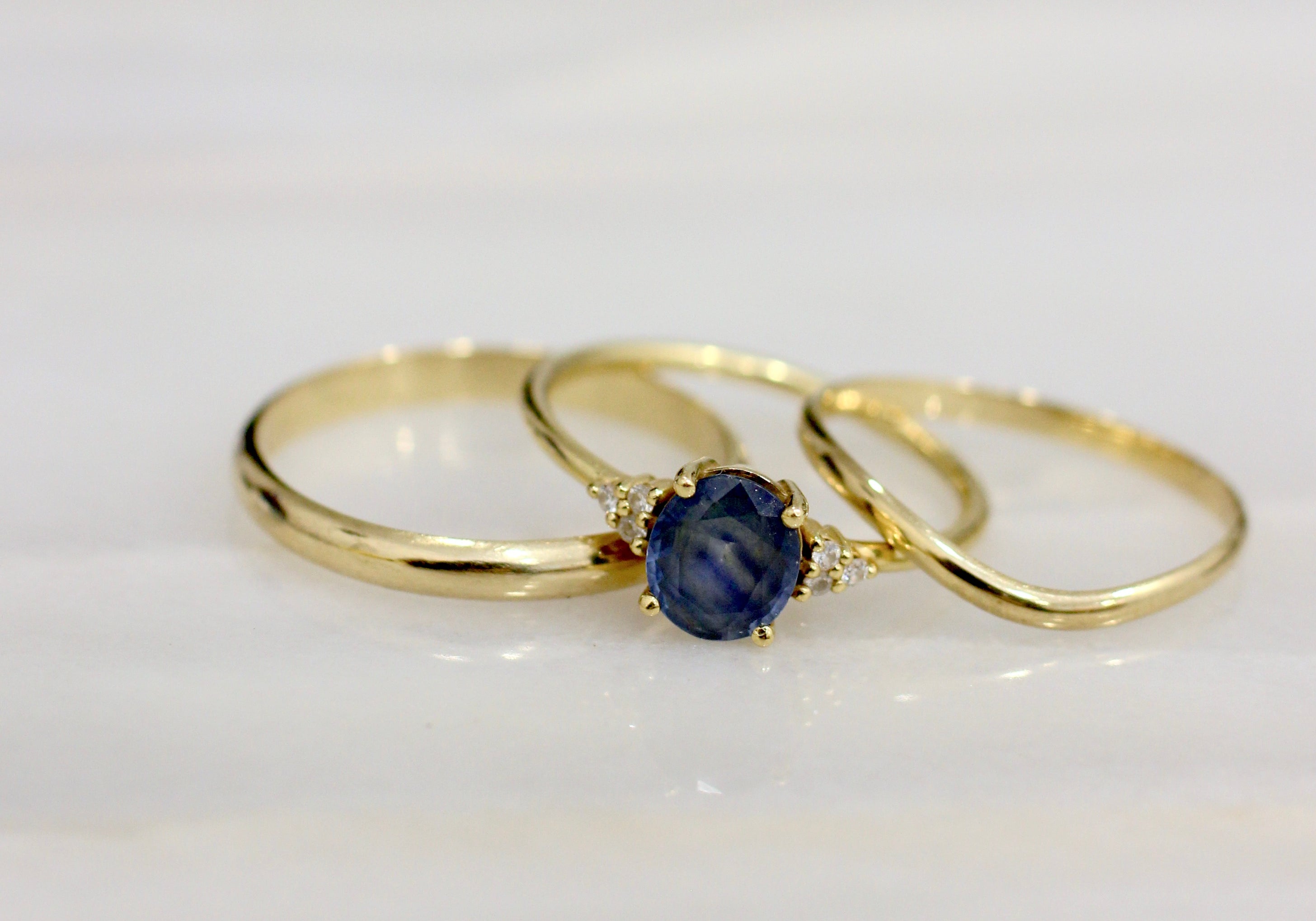 Two Yellow Gold wedding rings and an engagement ring with a Sapphire placed on a marble surface. 
