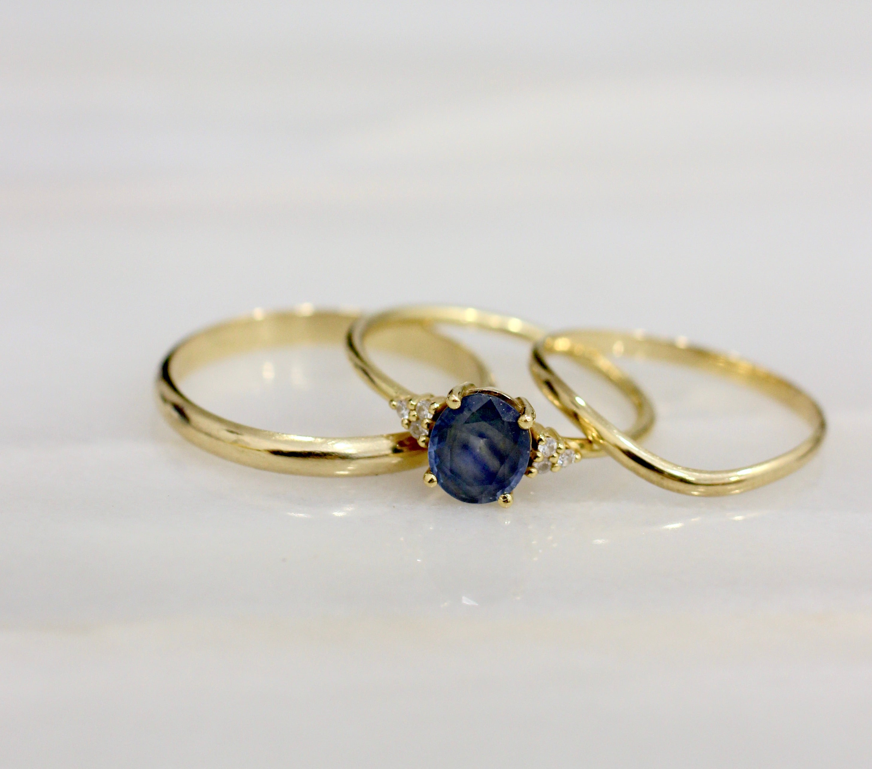 Two Yellow Gold wedding rings and an engagement ring with a Sapphire placed on a marble surface. 