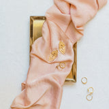 A pink silk scarf and gold rings on a vintage-inspired white surface, featuring The Guipure GP by Cremilde Bispo Jewellery.