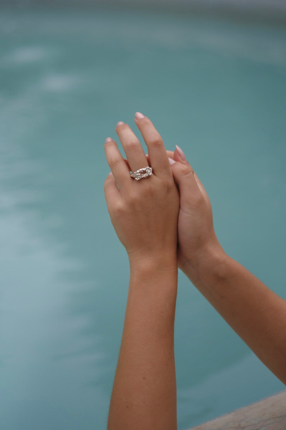 A handcrafted Glade ring adorned with floral foliage delicately held by a woman's hand near a serene pool in the glade, by Cremilde Bispo Jewellery.