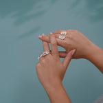 A woman's handcrafted hands adorned with a Cremilde Bispo Jewellery floral foliage jewelry collection, gently cradling The Glade ring by a pool.