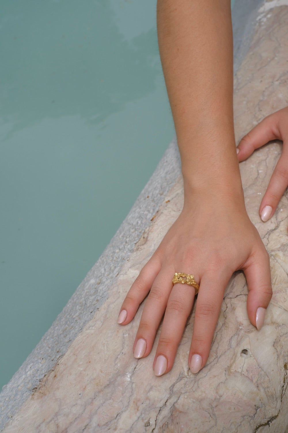 A woman's hand adorned with a handcrafted gold Glade ring from the Cremilde Bispo Jewellery collection, showcased elegantly next to a pool.