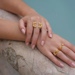 A woman's hand adorned with two gold rings from the Cremilde Bispo Jewellery Glade ring GP collection, glistening under the sunlight next to a pool.