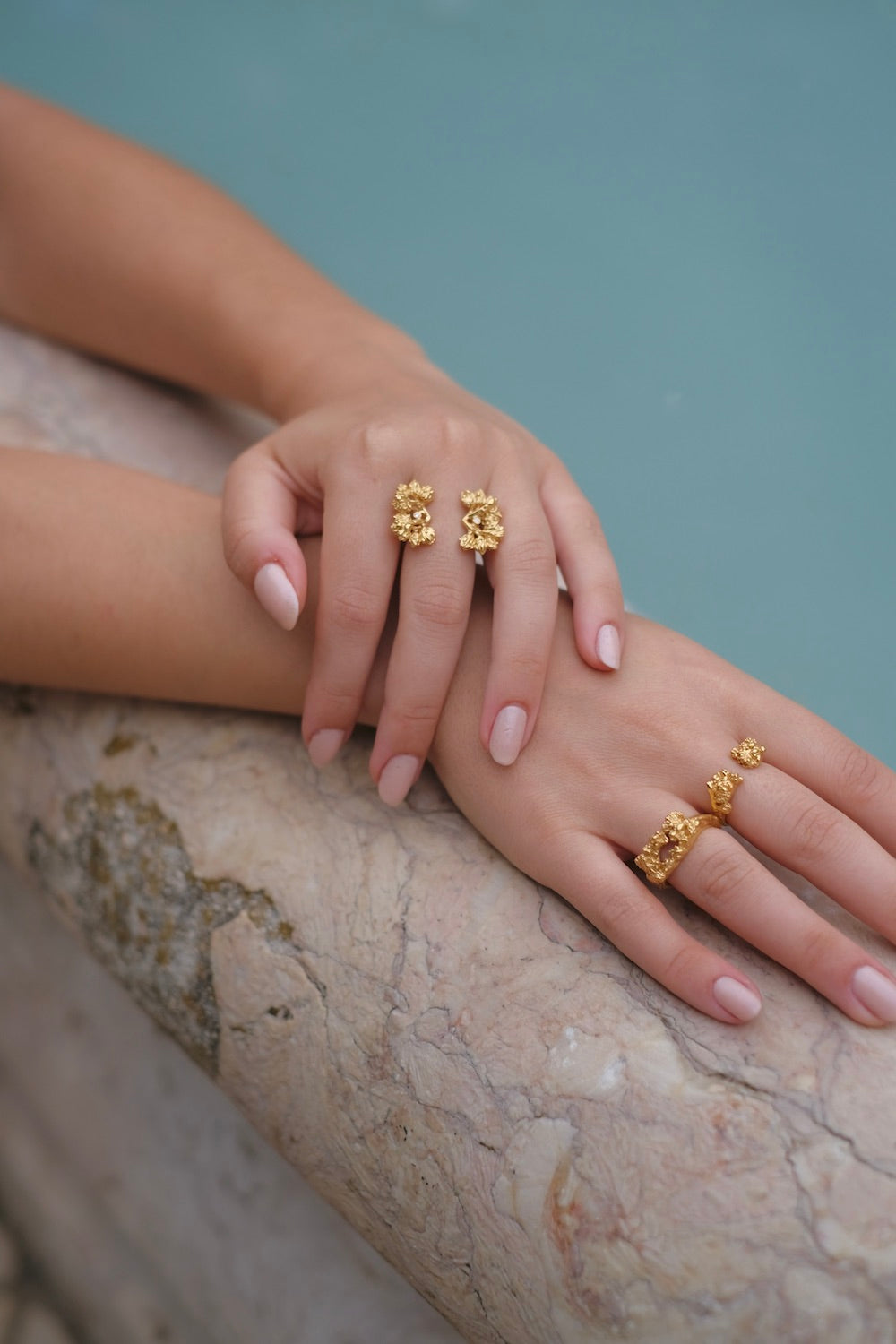 A woman's hand adorned with two gold rings from the Cremilde Bispo Jewellery Glade ring GP collection, glistening under the sunlight next to a pool.