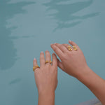 A woman's hands adorned with a Cremilde Bispo Jewellery's handcrafted jewelry collection, featuring The Glade ring GP, are elegantly displayed next to a pool.