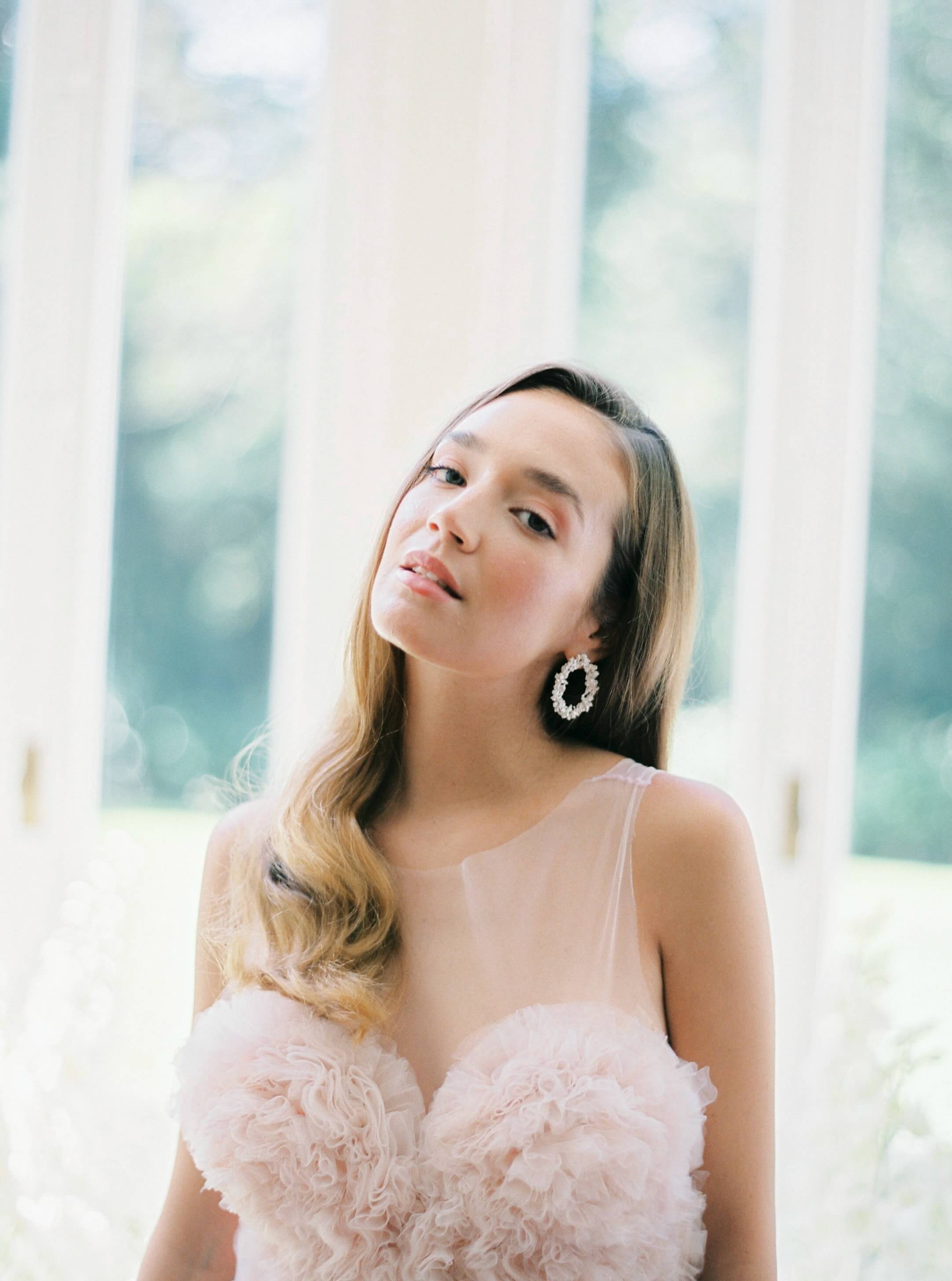 A bride in a pink dress gracefully posing in front of a window, showcasing the refined elegance of Nature's beauty, perfectly complemented by her Cremilde Bispo Jewellery Garden's Delight Earrings, in Sterling silver.