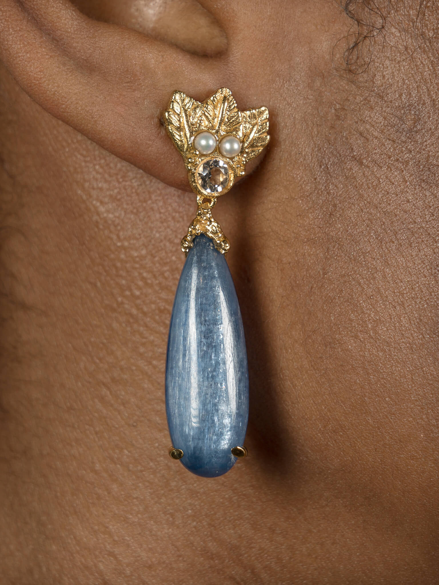 A woman's ear with a silver Cremilde Bispo Jewellery Muse III Kyanite earring, adorned with a blue teardrop, dangling from it.