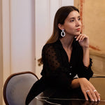 A sophisticated woman wearing the Cremilde Bispo Jewellery Garden's Delight statement earrings in sterling silver, seated at an opulently decorated table in a luxurious room.