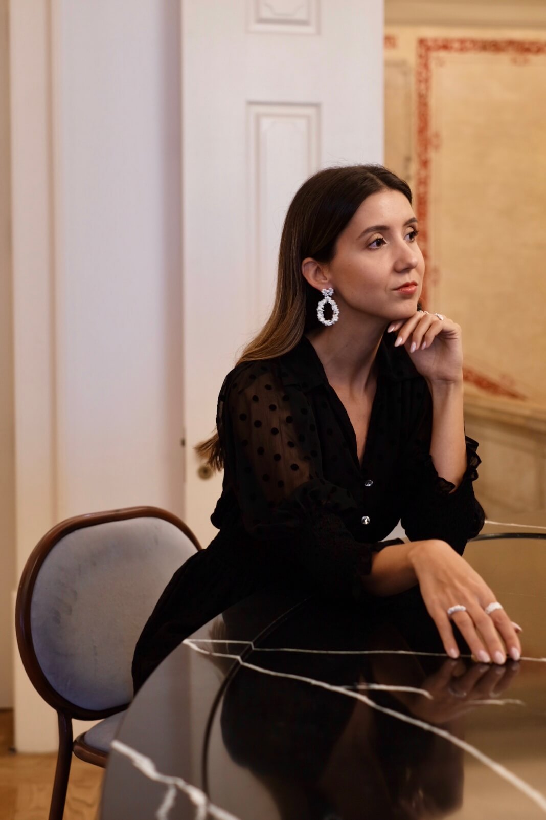 A sophisticated woman wearing the Cremilde Bispo Jewellery Garden's Delight statement earrings in sterling silver, seated at an opulently decorated table in a luxurious room.
