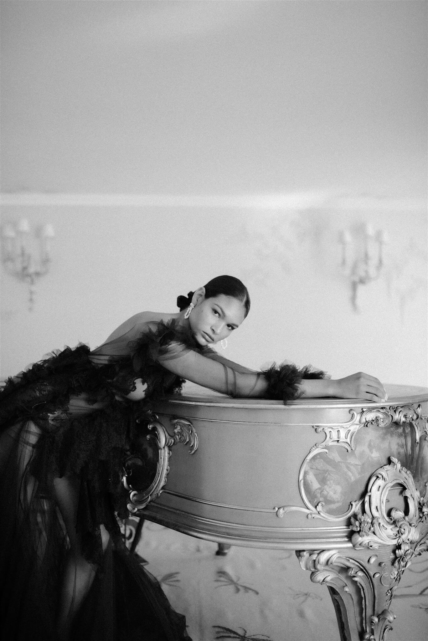 A black and white photo of a woman posing on a piano, showcasing opulence and wearing the Cremilde Bispo Jewellery earring set, The Garden's Delight II.