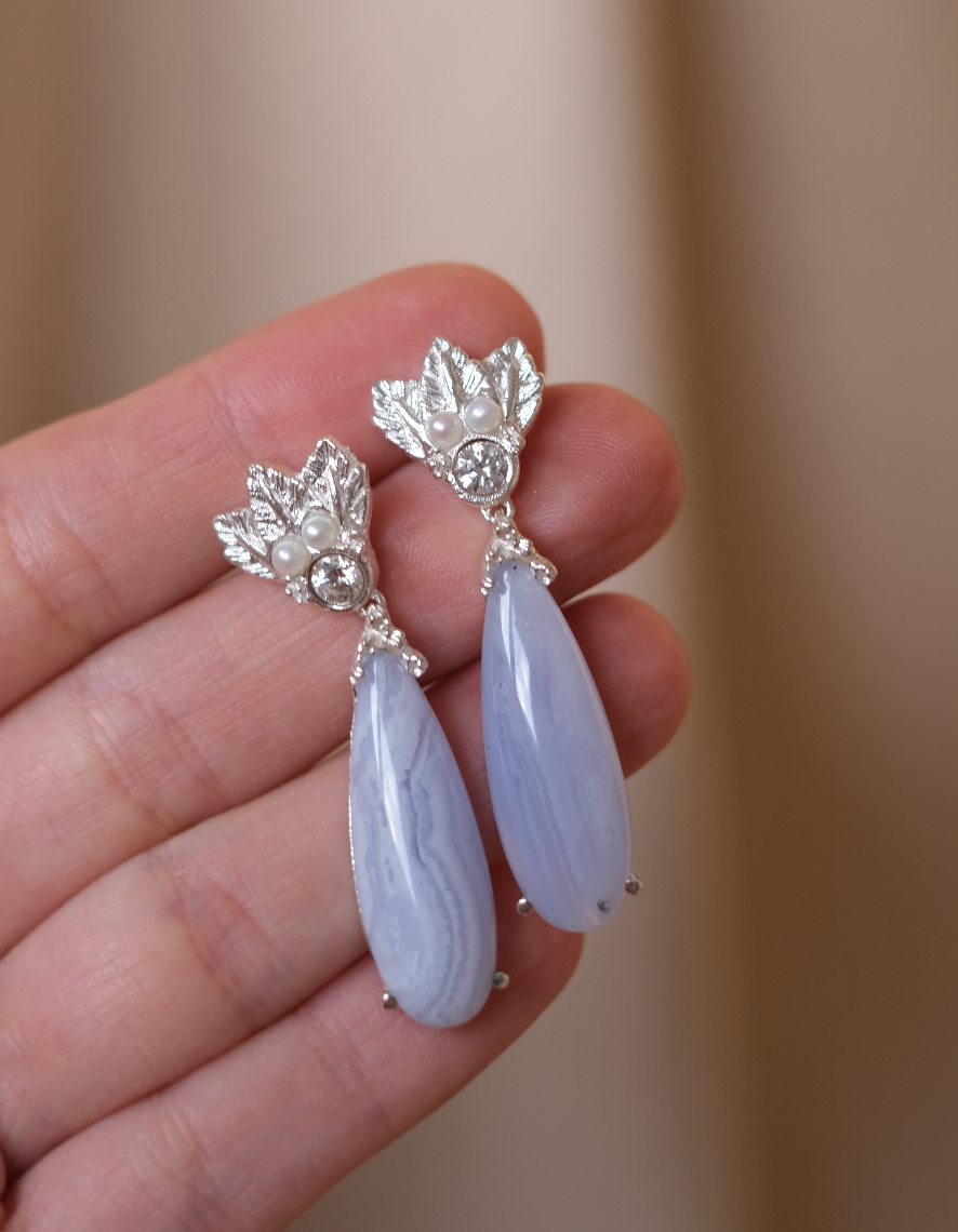 A hand holding a pair of The Muse III Chalcedony earrings, adorned with .925 silver accents by Cremilde Bispo Jewellery.