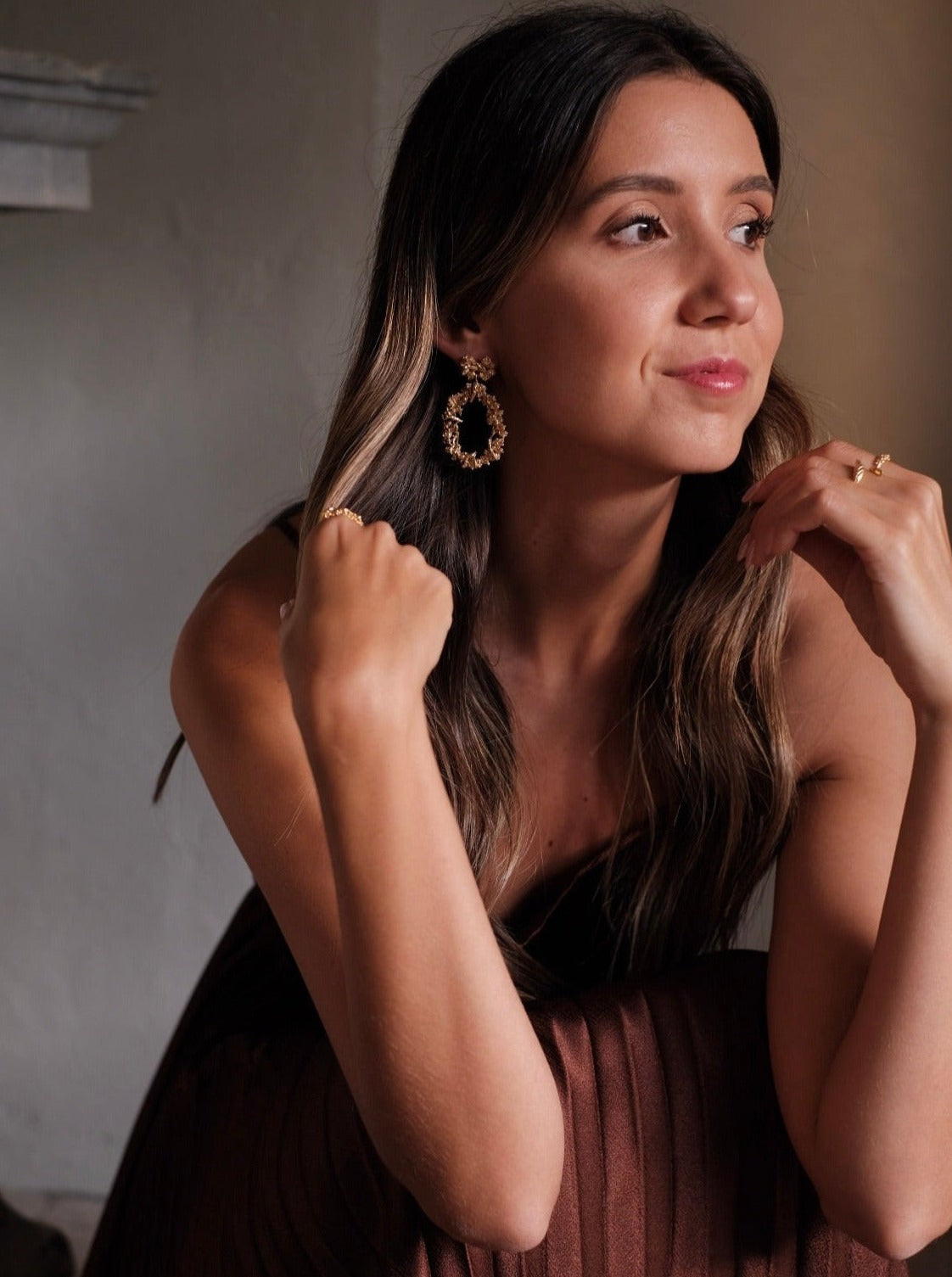 In a stunning display of opulence, a woman in a brown dress is posing for a photo. Her glamorous Cremilde Bispo Jewellery earring set, The Garden's Delight II GP, dazzles under the sunlight, enhancing her beauty as if she were the very