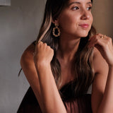 In a stunning display of opulence, a woman in a brown dress is posing for a photo. Her glamorous Cremilde Bispo Jewellery earring set, The Garden's Delight II GP, dazzles under the sunlight, enhancing her beauty as if she were the very