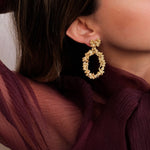 A woman wearing a burgundy blouse and gold earrings, exuding opulence with The Garden's Delight II GP earrings from Cremilde Bispo Jewellery.