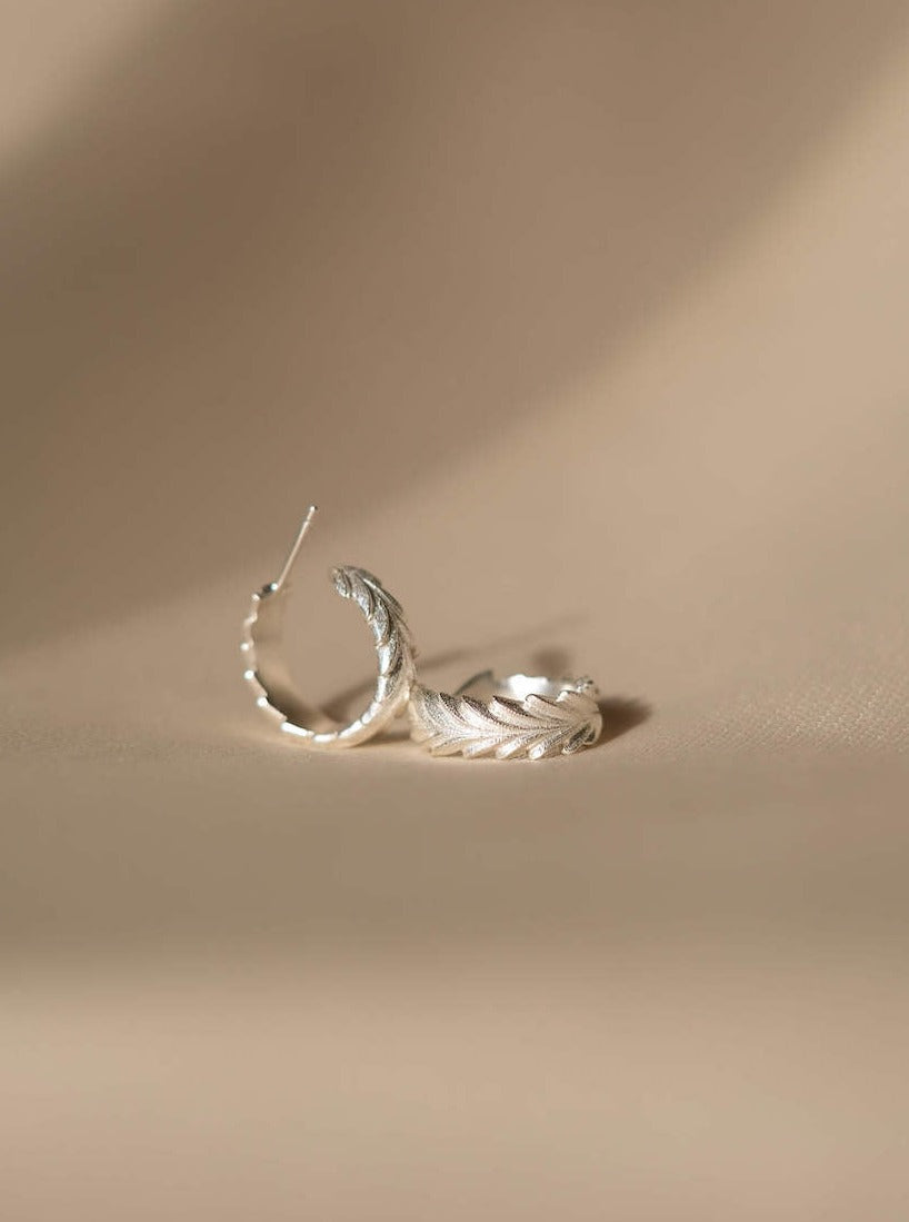 The Cremilde Bispo Jewellery Essential Leaf Hoops effortlessly exude maximum style as they rest delicately on a beige surface.