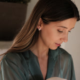 A woman in Lisbon is sitting on a bed, engrossed in her phone while wearing the stunning Cremilde Bispo Jewellery Essential Drops earrings from her jewelry collection.