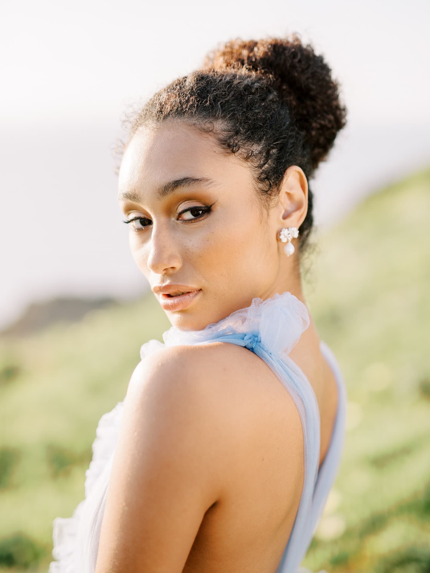 A stunning woman in a blue dress, adorned with Cremilde Bispo Jewellery's Pearly Forest earrings, posing elegantly in front of the vast ocean.